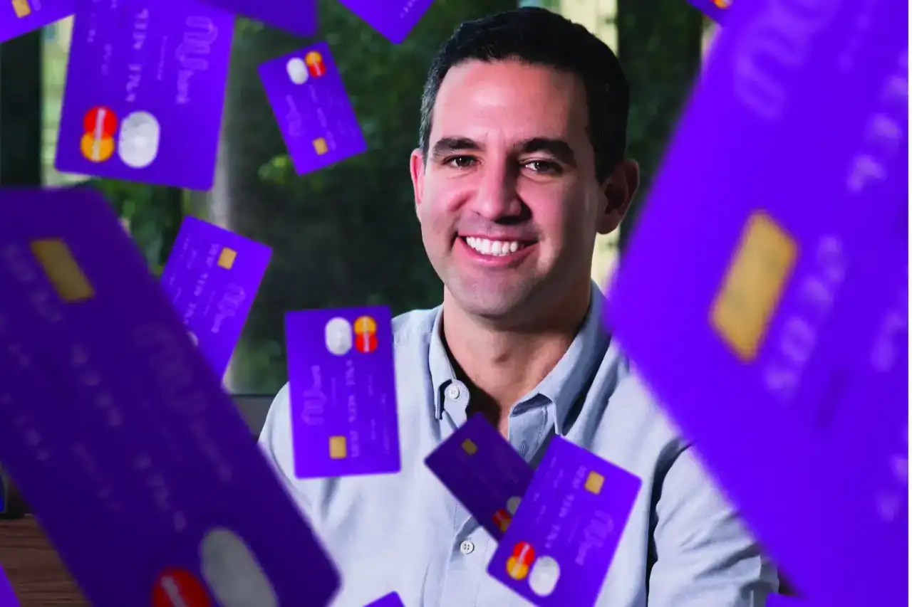 David Vélez, CEO do Nubank, sorrindo e olhando para a câmera em um cenário profissional que remete ao branding roxo da empresa, com um microfone à frente.