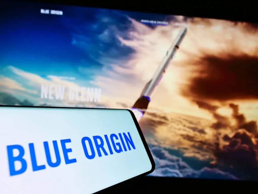 Logo da Blue Origin em um smartphone ao lado de uma imagem de um foguete, simbolizando a parceria na exploração lunar.