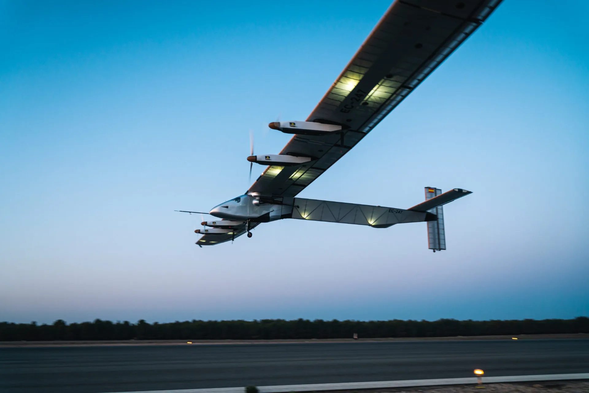 Drone Solar Impulse 2 em voo, com suas asas gigantescas repletas de painéis solares, sobrevoando uma paisagem.