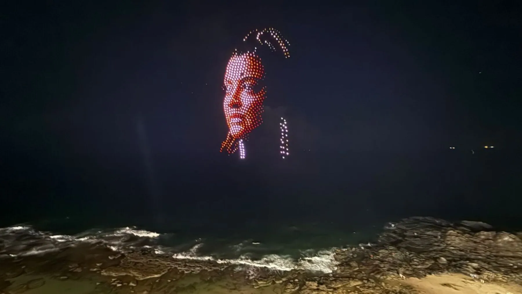 Imagem de 600 drones formando o rosto da cantora Preta Gil no céu noturno de Salvador, durante o Carnaval, com o circuito Barra-Ondina ao fundo.