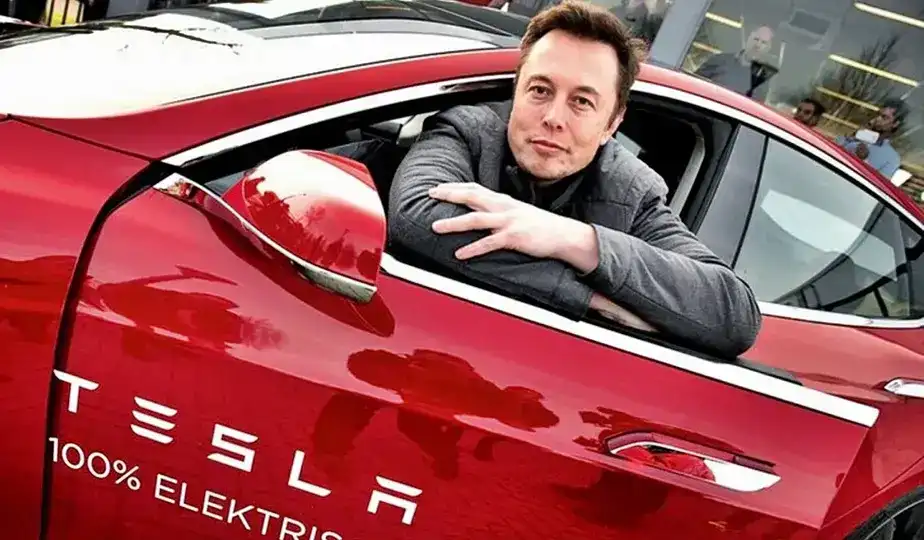 Elon Musk em um evento de tecnologia com o logotipo da Tesla ao fundo, representando a inovação em chips de IA.