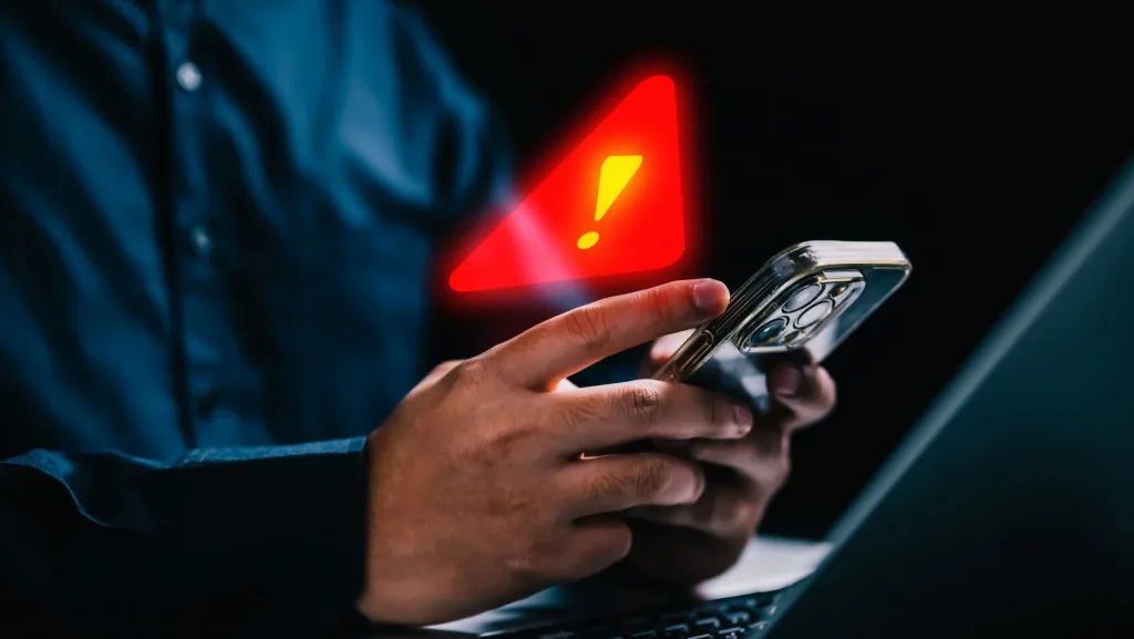 Imagem de uma pessoa segurando um smartphone com um símbolo de alerta de segurança na tela, ilustrando o perigo de golpes online e a necessidade de proteção.