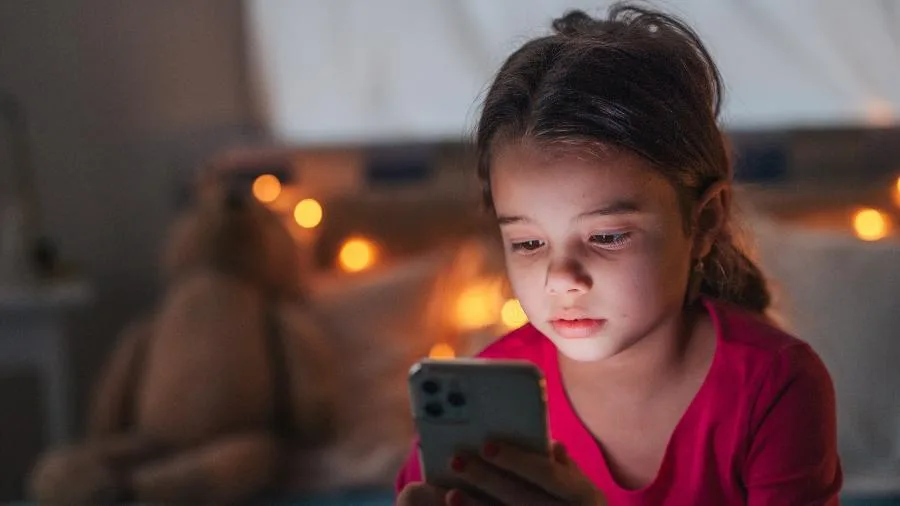 Criança sentada em uma cama, concentrada, olhando para a tela de um celular em um quarto escuro.