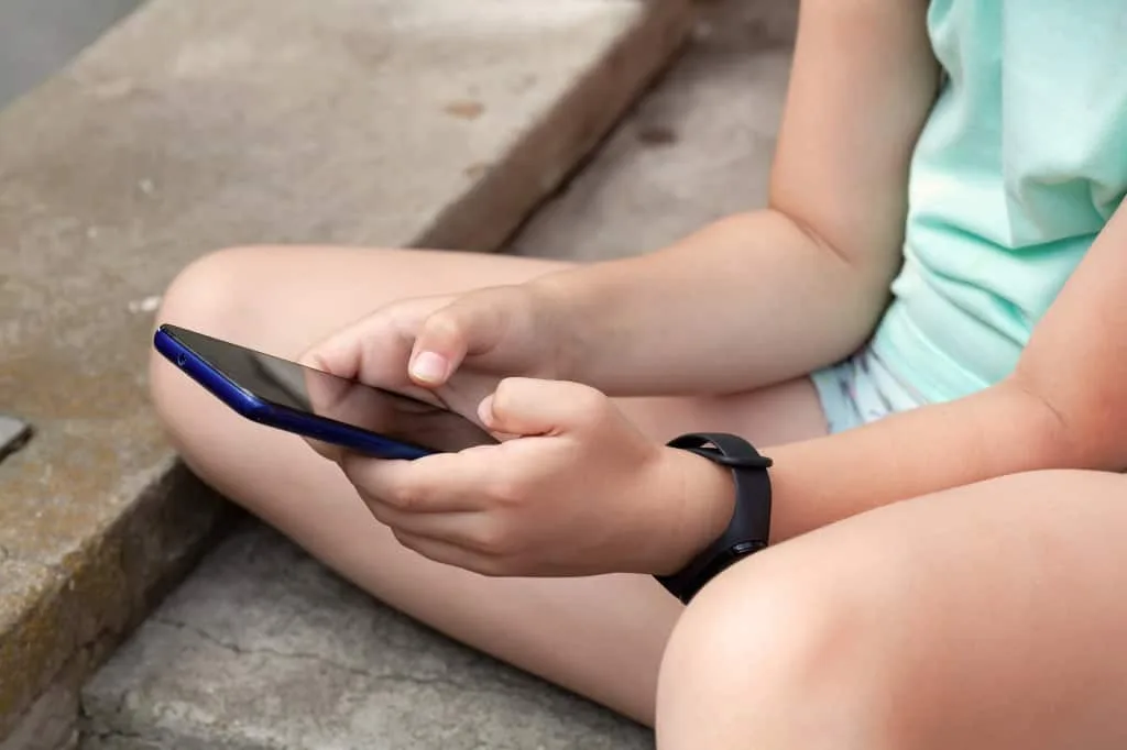 Criança concentrada mexendo em um celular, com foco nos dedos e na tela. A imagem ilustra a discussão sobre o impacto das redes sociais em usuários jovens.