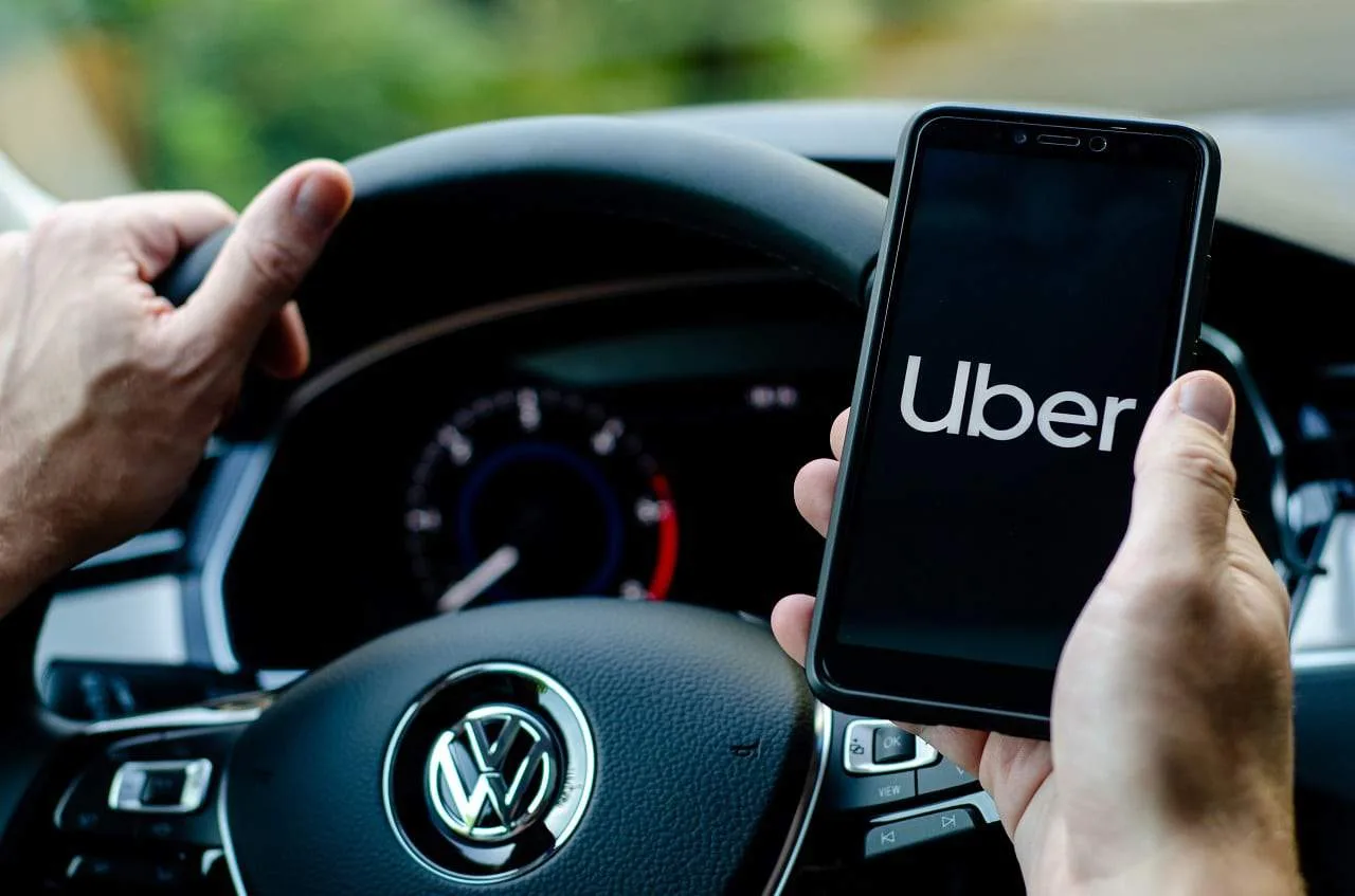 Logotipo da Uber em um celular com fundo de cidade movimentada, simbolizando o alcance global e a tecnologia de mobilidade. A imagem transmite a ideia de inovação e presença da marca em cenários urbanos.